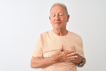 Senior grey-haired man wearing striped t-shirt standing over isolated white background smiling with hands on chest with closed eyes and grateful gesture on face. Health concept.
