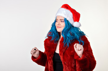 Christmas, fashion and youth - Beautiful young woman with blue hair in red fur coat and santa hat, looking at the camera and standing against a white background. Ready for party.