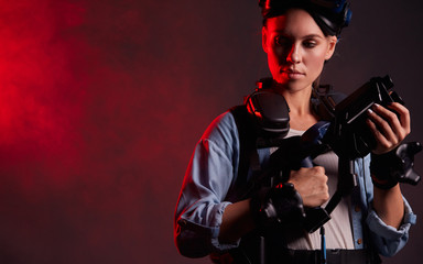 Attractive caucasian girl wearing special equipment, holding weapon for virtual game. Stand posing isolated over black background, red smoky neon backlit
