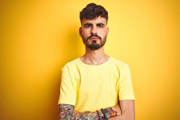 Young man with tattoo wearing t-shirt standing over isolated yellow background skeptic and nervous,...