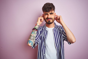 Obraz premium Young man with tattoo wearing striped shirt standing over isolated pink background covering ears with fingers with annoyed expression for the noise of loud music. Deaf concept.