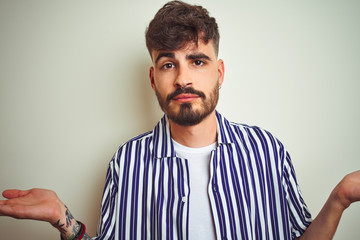 Young man with tattoo wearing striped shirt standing over isolated white background clueless and confused expression with arms and hands raised. Doubt concept.