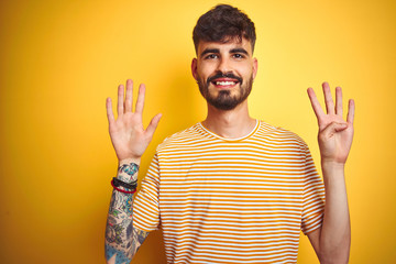 Young man with tattoo wearing striped t-shirt standing over isolated yellow background showing and...