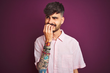 Young man with tattoo wearing summer pink shirt standing over isolated purple background looking stressed and nervous with hands on mouth biting nails. Anxiety problem.