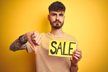 Young man with tattoo holding sale banner standing over isolated yellow background with angry face,...
