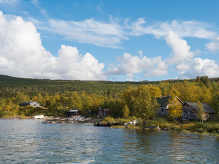 Fototapeta na wymiar View on sami hut village in Saltoluokta from river Lulealven with green mountains, yellow birch tree and blue sky white clouds, golden hour light