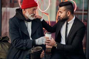 merciful businessman sit next to tramp in red hat and coat, rich man in black tuxedo. Vagrant get coffee from businessman