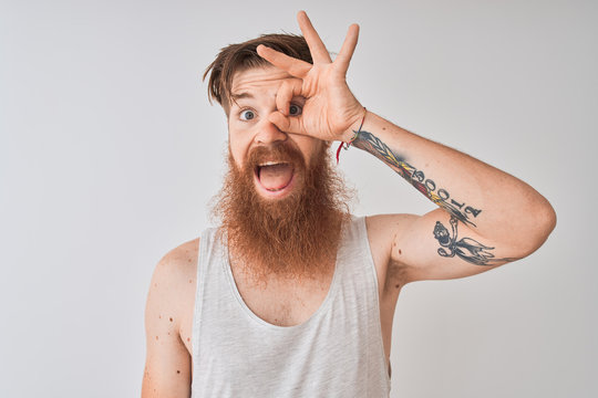Young redhead irish man wearing t-shirt standing over isolated grey background doing ok gesture with hand smiling, eye looking through fingers with happy face.
