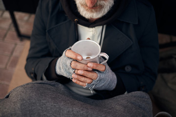 Homeless man's hands holding half empty mug with some coins at bottom. Hands in grey gloves....