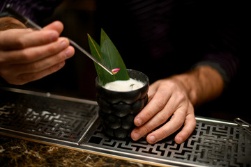 Male bartender decorated a glass with cocktail drink with a green tropical leaf and dried rose buds with tweezers