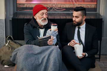Young beardy man in suit sitting with beggar on floor on street and give cup of coffee. Different segments of society, social inequality