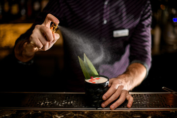 Bartender spaying on a black glass with cocktail decorated with a green tropical leaf, dried rose bud and essence drops