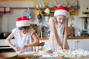 Children are cooking Christmas cookies from dough in cozy home kitchen. Cute kids in santa hats preparation holiday family dinner. Girls bake Xmas biscuits. Lifestyle moments. Children chef concept