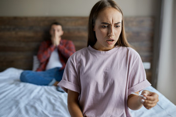 Frustrated young pretty girl knitting her brows with horrified expression, stares at pregnancy test, gets positive result, looks away with shocked emotions, blurred background, close up
