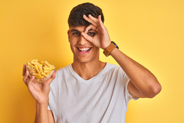 Young indian man holding bowl with dry pasta standing over isolated yellow background with happy face smiling doing ok sign with hand on eye looking through fingers