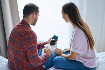 Charming young couple spending holidays together at home, staring at cell phone screen, looking for number, drinking hot tea, holding white cup, sitting on sheets with backs towards camera