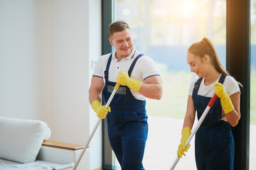 Staff of cleaning service work together, washing floor with mop. Wearing special clothes for cleaning. Panoramic window background