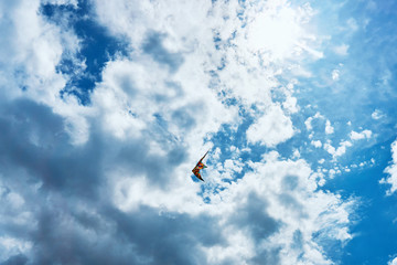 A large kite floats high in the sky among the clouds against the blue sky and the bright summer sun and it is controlled by two white ropes going to the ground
