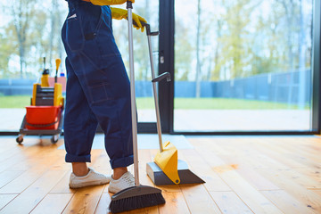 Side view on young cleaner sweeping the floor in living room, panoramic window background. Cleaning...