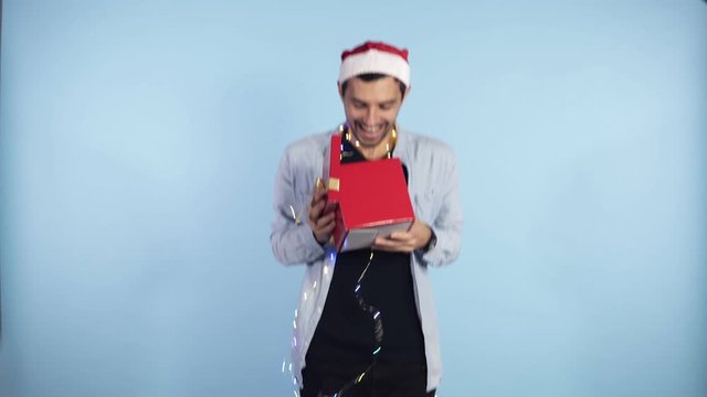 Positive, young guy in Christmas hat and garland on neck recieves surprise from from the side female hands - being in good festive mood, isolated over blue background. People, surprisment and presents