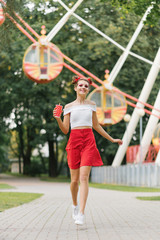 A young woman with bright make-up holds a red paper cup in her hands and runs through an amusement park. She is smiling and happy.