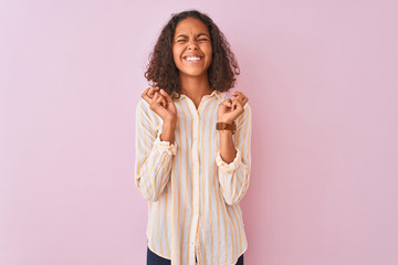 Young brazilian woman wearing striped shirt standing over isolated pink background gesturing finger crossed smiling with hope and eyes closed. Luck and superstitious concept.