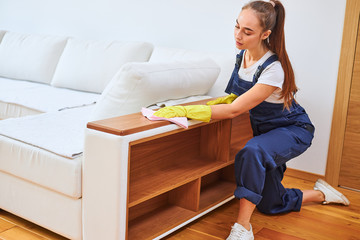 Youthful woman wearing yellow rubber gloves, blue uniform, hair tied in a ponytail, wiping dust off...