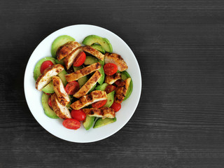 Grilled chicken salad with avocado and tomatoes on a white plate with black gray wood table background. Copy space is on the right side. Top view photo. 