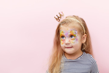 Pretty exciting smiling blond little child girl of 3 years with a bright face painting on pink background. Fun emotions, happy childhood