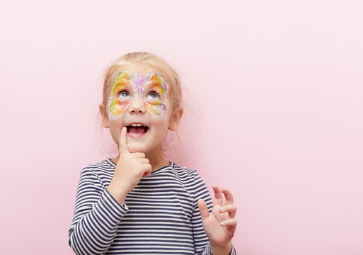 Pretty exciting smiling blond little child girl of 3 years with a bright face painting on pink background. Fun emotions, happy childhood. Holds a finger near his mouth, thinks and looks up