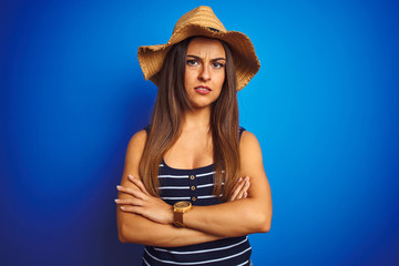 Young beautiful woman wearing striped t-shirt and summer hat over isolated blue background skeptic and nervous, disapproving expression on face with crossed arms. Negative person.