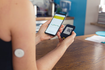 Woman comparing result of glucose level check on the traditional glucometer and on the mobile phone reading from modern technology remote sensor mounted on her forehand