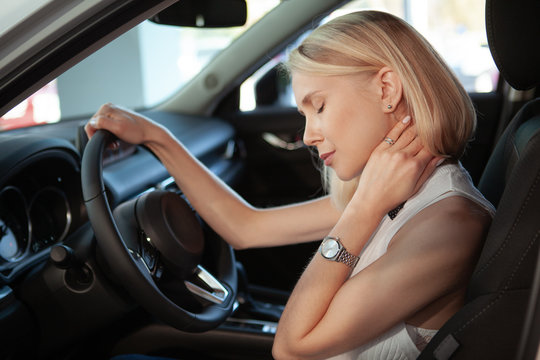Beautiful blond haired woman rubbing her aching neck, sitting in her car. Female driver rubbing her neck, tired from driving