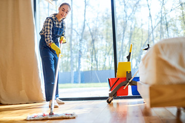 Process of mopping floor by young female in blue uniform, trolley of detergents next to her