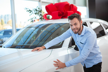 Charming handsome man smiling to the camera, touching beautiful new car with red bow on the roof at...