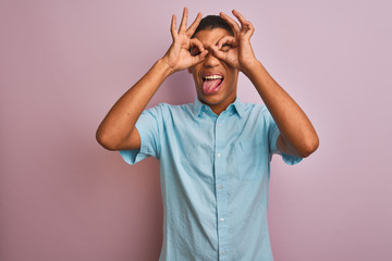 Young handsome arab man wearing blue shirt standing over isolated pink background doing ok gesture like binoculars sticking tongue out, eyes looking through fingers. Crazy expression.
