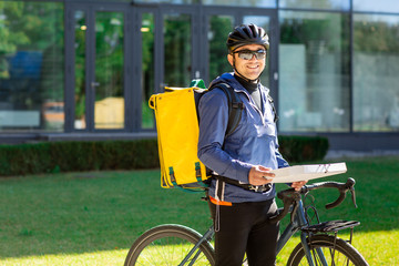 Fototapeta na wymiar Portrait of bicycle courier with yellow bag and bike. Man in helmet and glasses holding pizza box