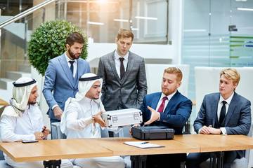 Sheikh in white suit give white case with money in barter for getting new modern technology by caucasians with black case. Caucasian men in tuxedo. Sit on table, office background