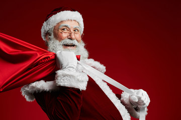 Waist up portrait of smiling Santa Claus holding bag with Christmas presents over red background,...