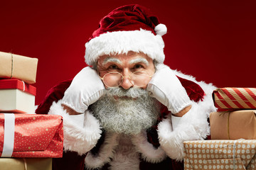 Close up portrait of fairytale Santa looking at camera while posing with Christmas presents over red background