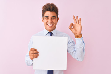 Young handsome businessman holding banner standing over isolated pink background doing ok sign with fingers, excellent symbol
