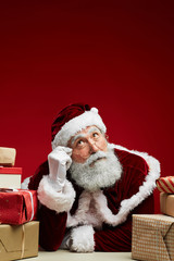Portrait of fairytale Santa Claus looking up and thinking while posing with Christmas presents over red background