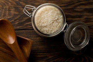 White uncooked, raw long grain rice in glass storage jar on rustic brown wooden table top view flat lay from above