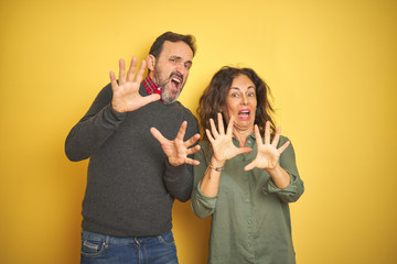 Beautiful middle age couple over isolated yellow background afraid and terrified with fear expression stop gesture with hands, shouting in shock. Panic concept.
