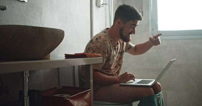 Bearded young man working on laptop computer sitting on toilet in the bathroom. Cheerful business guy cheering up checking good news on notebook.