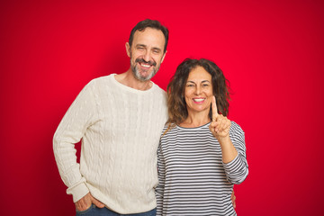 Beautiful middle age couple wearing winter sweater over isolated red background showing and pointing up with finger number one while smiling confident and happy.
