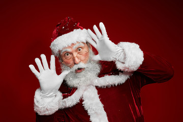 Waist up portrait of classic Santa Claus making funny faces while posing against red background in studio, copy space