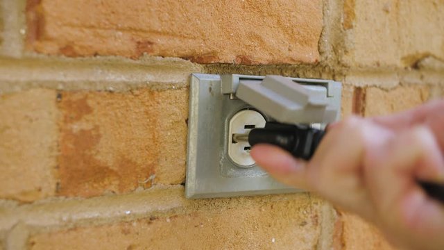 Man plugging in a power cord and then unplugging it from an outdoor electrical outlet. Dual covered outdoor power sockets on brick wall. Black extension cord plug inserted into a wall socket.