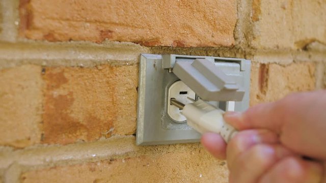 Man plugging in a power cord and then unplugging it from an outdoor electrical outlet. Dual covered outdoor power sockets on brick wall. White extension cord plug inserted into a wall socket.