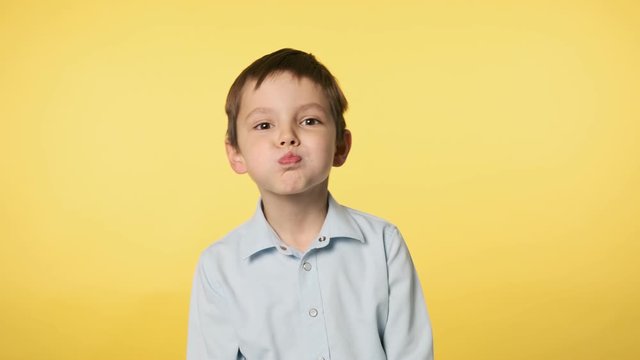 Funny little boy playfully making faces looking at camera. Positive emotion. Caucasian kid boy in blue shirt doing silly funny face puffing out cheeks at yellow background. Medium shot in 4k, UHD